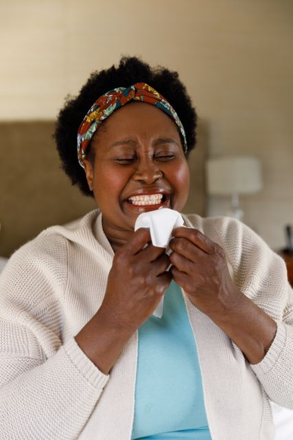 Close up of an old african-american woman with a grinning while holding a tissue close to her face. behind her is a bed and a lamp.