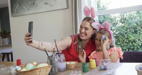 Mother and daughter wearing bunny ears painting easter eggs and taking playful selfie