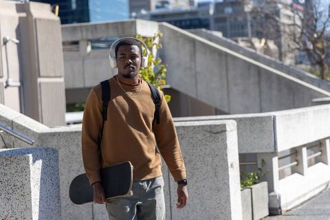 African American man walking with skateboard and headphones through urban concrete plaza