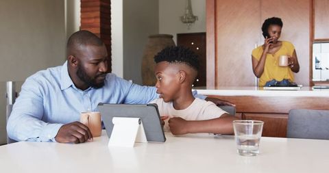 Caring African American father helping son with tablet at kitchen table, mother on phone