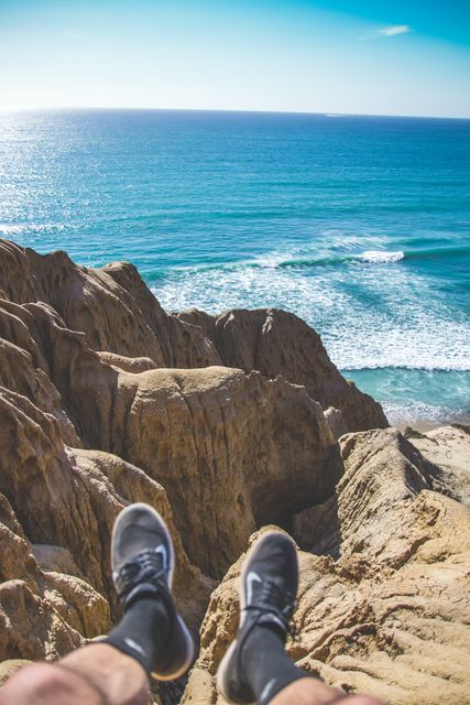 This image depicts a person sitting at the edge of cliffs with their feet dangling and a breathtaking view of the ocean in the background. Ideal for travel inspiration, nature photography, adventure blogs, and promotional material for outdoor activities.