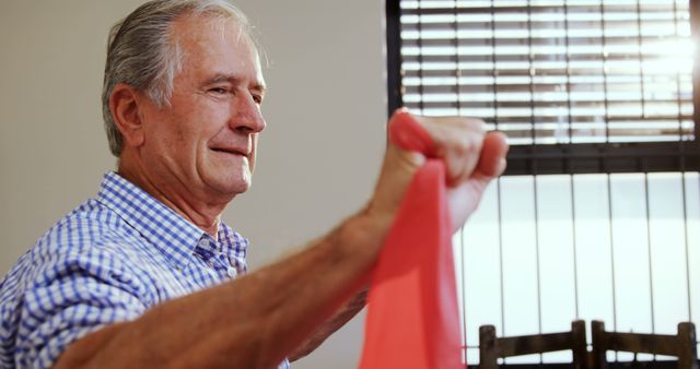 Senior man using resistance band for exercise at home, in a well-lit room with window blinds in background. This image can be used for health and wellness promotions, fitness campaigns, or articles on staying active in senior years. Ideal for illustrating home workout routines and promoting active lifestyles among older adults.