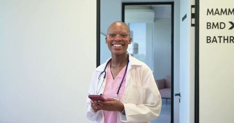 African clinician smiling while holding smartphone wearing stethoscope and pink scrubs