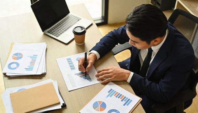 Business analyst annotating financial charts at desk with laptop and coffee