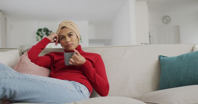 Image of biracial woman in hijab sitting on sofa and drinking coffee. Lifestyle, spending free time at home with technology concept.