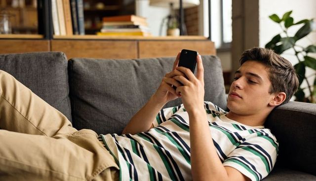 Teen relaxing on sofa using smartphone, casual home leisure with books and indoor plant