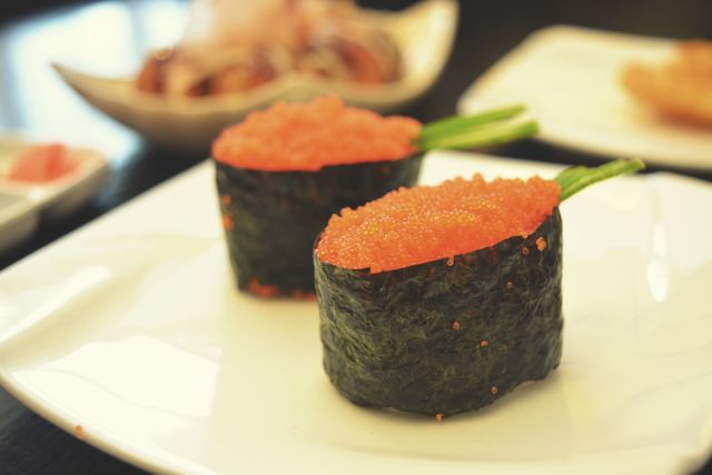 Gunkan-maki sushi with orange fish roe and wrapped in seaweed, presented on a white plate. Ideal for use in articles about Japanese cuisine or food blogs. Suitable for menu design, culinary magazines, or promotional materials for Japanese restaurants.