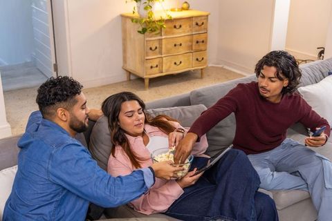 Three friends sharing popcorn on couch while streaming movie on tablet during cozy night
