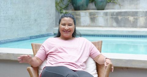 Mature african american woman sitting by backyard pool, smiling and relaxing in wicker chair