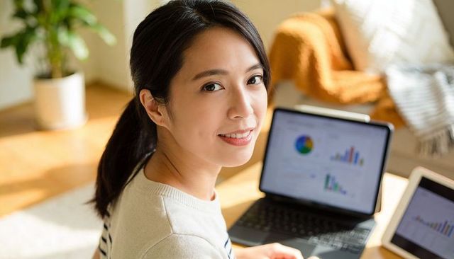Asian woman reviewing analytics on laptop and tablet in cozy living room workspace