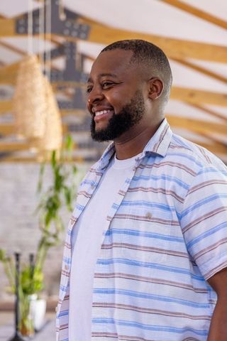 Smiling bearded man standing in cozy coffeehouse wearing striped shirt, pendant lamps, greenery
