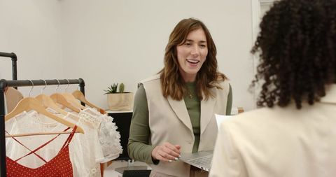 Boutique salesperson helping customer with laptop at clothing counter, dresses on rack