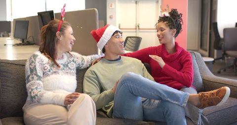 Diverse coworkers sharing holiday cheer on office lounge sofa wearing Santa hat, reindeer headband