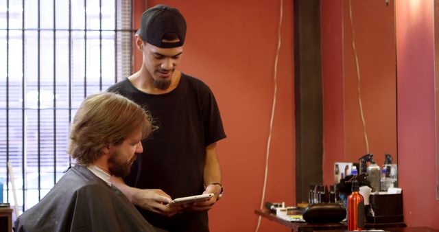 Barber showing client haircut options using a tablet in a modern barbershop. Suitable for depicting themes of personal grooming, customer service, modern barbershops, hairstyling consultations, and trendy hairstyles.