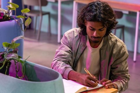 Young indian man writing in notebook at modern coworking lounge with gold pen and greenery