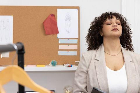 Fashion designer taking calm breath in studio with corkboard, fabric swatches, tablet