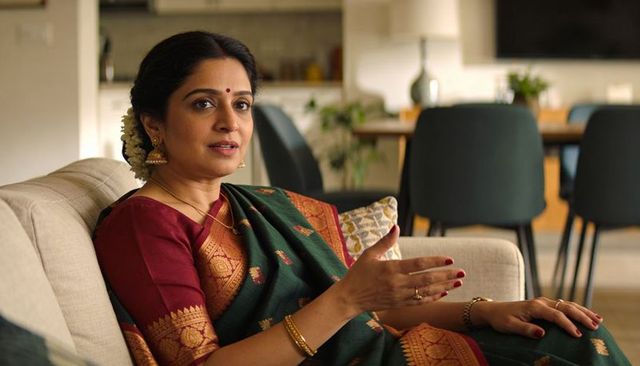 Indian woman in silk saree gesturing and speaking on sofa, maroon green tones with gold jewelry