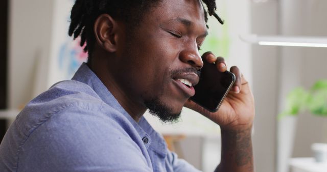 The image shows a young man talking on a smartphone in a home office while smiling, suggesting positive conversation. Ideal for illustrating concepts related to remote work, technology in everyday life, and modern communication. Can be used in marketing materials, blog posts about work-from-home setups, and advertisements for communication services.