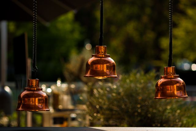Outdoor scene with a focus on three vintage-style copper lamps hanging on spiraled cords. Sunlight highlighting warm copper tones, creating a cozy ambiance. Ideal for articles or advertisements related to outdoor decor, patio design, nightlife venues, antique lighting styles, and events. Useful for marketing materials, blogs, and websites showcasing rustic or vintage-themed decor.