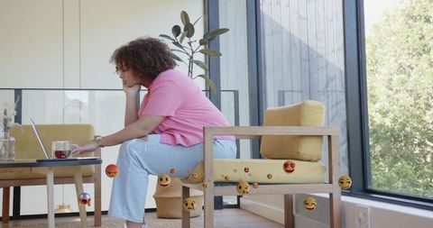 Woman working remotely on laptop in cozy sunlit living room with floating emoji reactions