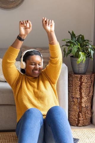 African american woman listening to music with headphones raising arms in cozy living room