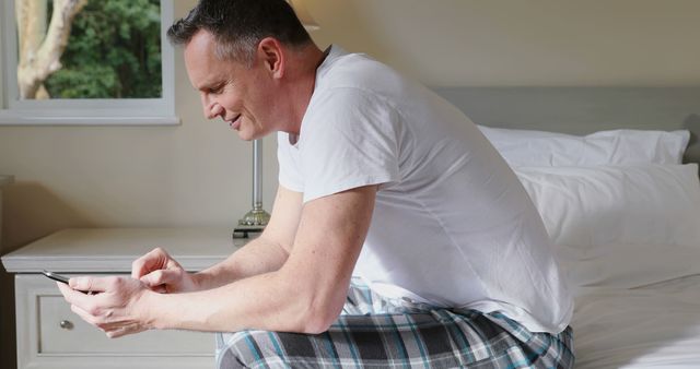 A middle-aged man sits on a bed in a modern bedroom while using a smartphone. He wears casual pajamas and appears comfortable and engaged with his device. This image is perfect for illustrating themes such as home life, technology use in daily routines, relaxation, and modern lifestyle.