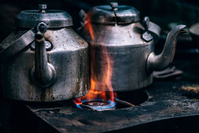 Shows old metal teapots boiling water on a gas stove in outdoor cafe. Suitable for illustrating traditional cooking methods, vintage cookware, street food preparation, or rustic kitchen settings. Can be used in articles about cultural heritage, travel, and culinary traditions.