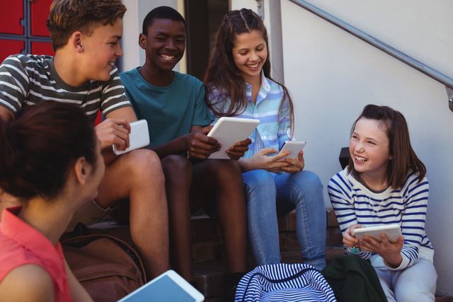 Group of diverse students sitting on a school staircase, engaging with mobile phones and tablets. They are smiling and appear to be enjoying their time together. This image can be used for educational content, technology in education, student life, and social interaction themes.