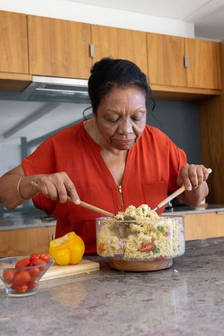 Senior african american woman mixing colorful pasta salad in modern kitchen