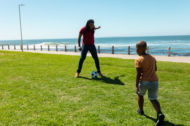 Father and son enjoying a sunny day playing soccer on a grassy area near the beach. Perfect for illustrating family bonding, outdoor activities, and leisure time. Ideal for use in advertisements, blogs, and articles about family life, sports, and healthy living.