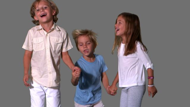 Group of cheerful kids joyfully holding hands and laughing while jumping indoors against grey background. Perfect for concepts of friendship, joy, childhood, and playful moments. Great for promotional materials, family-oriented products, and educational content.