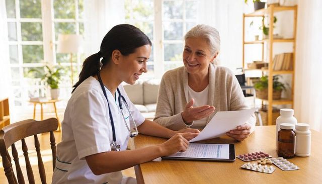Home care nurse reviewing medication plan with senior woman using tablet at kitchen table