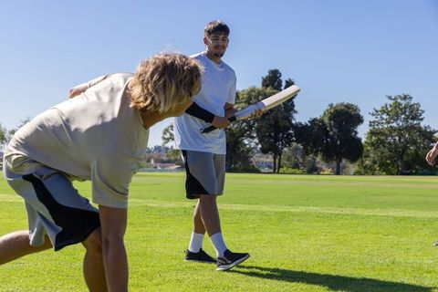 Young diverse friends playing cricket on park field, batting and fielding, outdoor sport