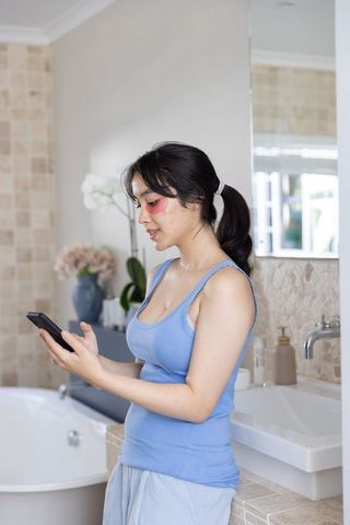 Asian woman applying under-eye patches while checking smartphone in modern bathroom