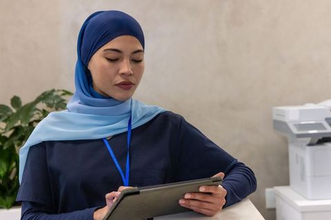 Healthcare receptionist wearing blue hijab standing at clinic desk holding tablet