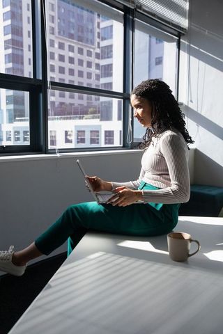 African-american woman sitting on desk using laptop in sunlit modern office, teal pants