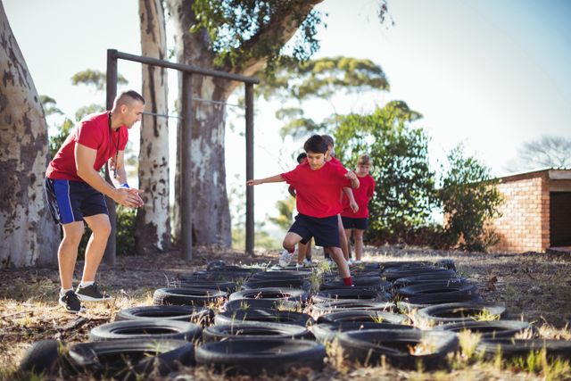 Trainer guiding kids through a tire obstacle course in an outdoor boot camp. Ideal for use in articles or advertisements related to fitness training for children, outdoor activities, summer camps, physical education programs, and teamwork exercises.