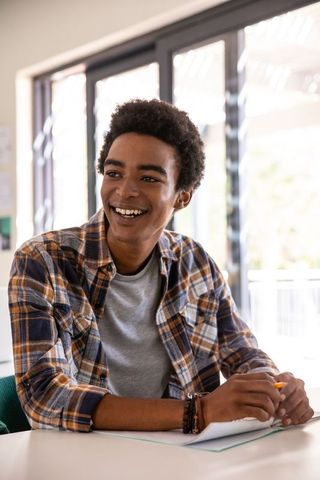 African American Teen Smiling While Studying at Desk Wearing Plaid Shirt in Classroom