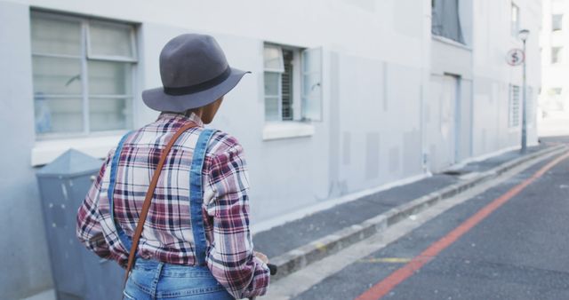 Woman walking down urban street wearing hat, plaid shirt, and jeans. She is in casual attire suitable for everyday wear. This can be used for urban lifestyle, fashion, and street photography themes. Great for illustrating modern living, casual fashion, and youthful trends.