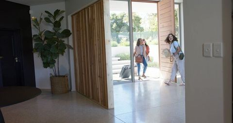 Three women entering sunlit modern foyer carrying suitcase, bags and sun hat on arrival