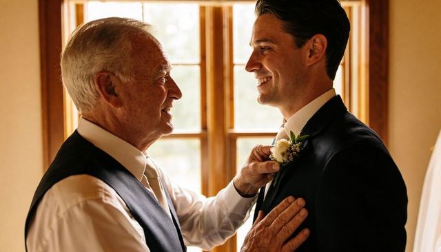 Elder man fastening boutonniere on smiling groom near sunlit wooden window