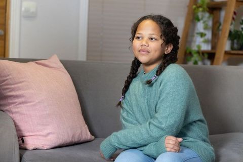 Calm preteen girl sitting on gray sofa wearing teal sweater and braided pigtails