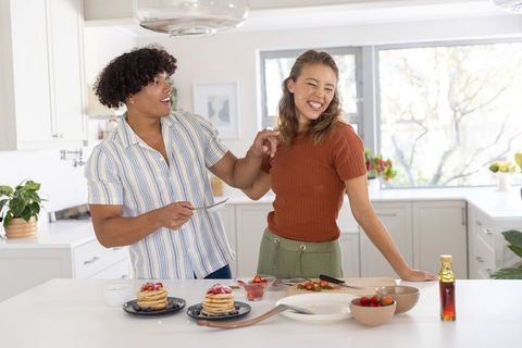 Young couple laughing and plating pancakes on bright modern kitchen island during morning breakfast
