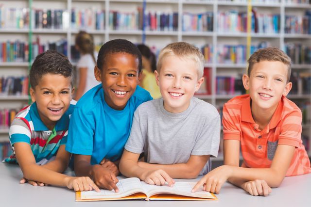 Four boys of different ethnic backgrounds are reading a book together in a school library. They are smiling and appear engaged, showcasing teamwork and friendship. This image can be used for educational materials, school websites, library promotions, and articles on diversity and learning.