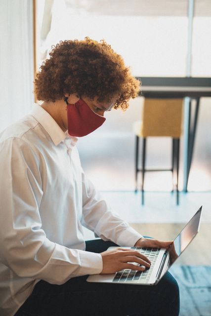 Biracial businessman wearing a face mask is working on his laptop in a hotel lobby. This image is ideal for illustrating concepts related to business travel during the COVID-19 pandemic, remote work, safety measures, and professional environments. It can be used in articles, blogs, and promotional materials related to business, travel, health and safety protocols, and modern work practices.