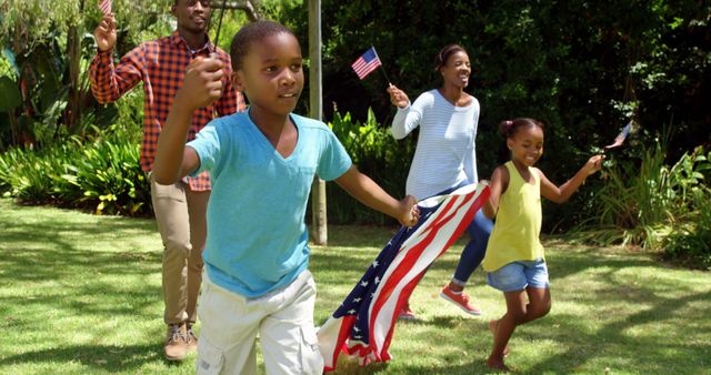 African American family including parents and children celebrating the 4th of July outdoors in a park. They are holding American flags and seem to be having fun together in the grassy area. This image portrays patriotism, family bonding, and summertime activities. It can be used for promotions related to holiday celebrations, family gatherings, patriotic events, and summer outings.