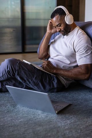 Mature african man studying at home with headphones writing in notebook next to laptop