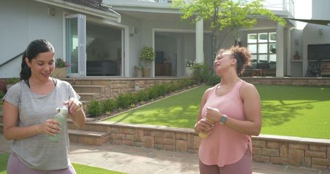 Laughing diverse women holding reusable water bottles on backyard patio after workout