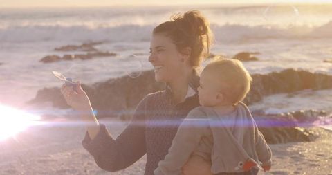 Mother holding baby blowing bubbles on sandy beach at sunset, golden hour bonding