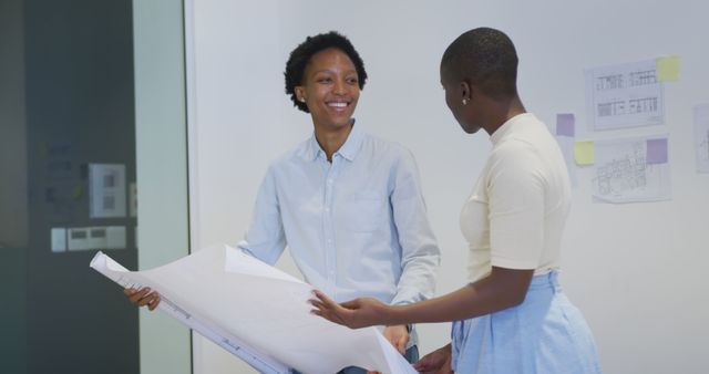 Two female architects engaging in a collaborative discussion while reviewing blueprints. Ideal for using in subjects related to architecture, teamwork, professional collaboration, or workplace diversity.